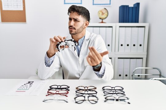 Young optician man holding optometry glasses beckoning come here gesture with hand inviting welcoming happy and smiling