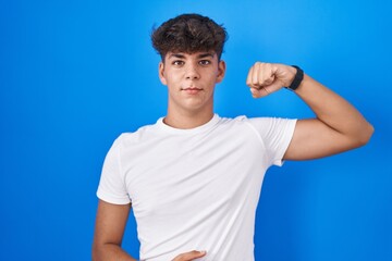 Hispanic teenager standing over blue background strong person showing arm muscle, confident and...