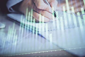 A woman hands writing information about stock market in notepad. Forex chart holograms in front. Concept of research. Multi exposure