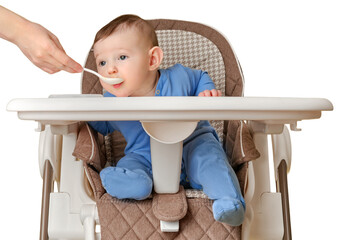 Mother feeding toddler baby from a spoon on a high chair for feeding children, isolated on a white background. Child boy at age of six months eats applesauce while sitting on a baby chair, copy space