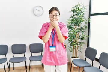 Young asian nurse woman at medical waiting room shocked covering mouth with hands for mistake. secret concept.