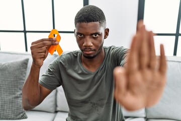 Young african american man holding awareness orange ribbon sitting on the sofa with open hand doing...