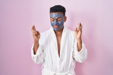 Young hispanic man wearing beauty face mask and bath robe gesturing finger crossed smiling with hope and eyes closed. luck and superstitious concept.