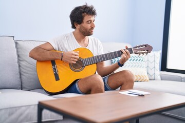 Young hispanic man playing classical guitar sitting on sofa at home
