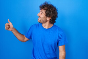 Hispanic young man standing over blue background looking proud, smiling doing thumbs up gesture to the side