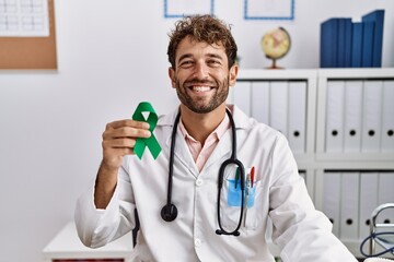 Young hispanic doctor man holding support green ribbon at clinic looking positive and happy...