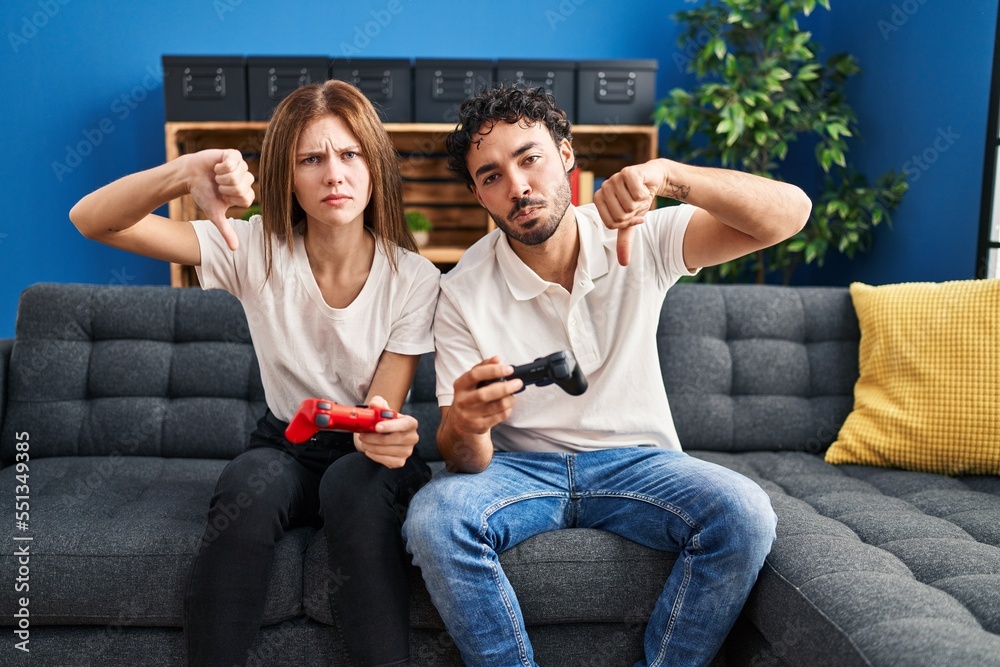 Sticker young couple playing video game holding controller at home with angry face, negative sign showing di