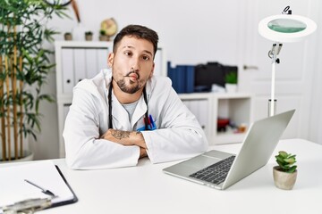 Young doctor working at the clinic using computer laptop making fish face with lips, crazy and comical gesture. funny expression.
