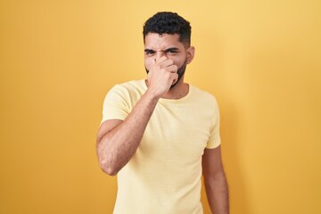 Hispanic man with beard standing over yellow background smelling something stinky and disgusting,...