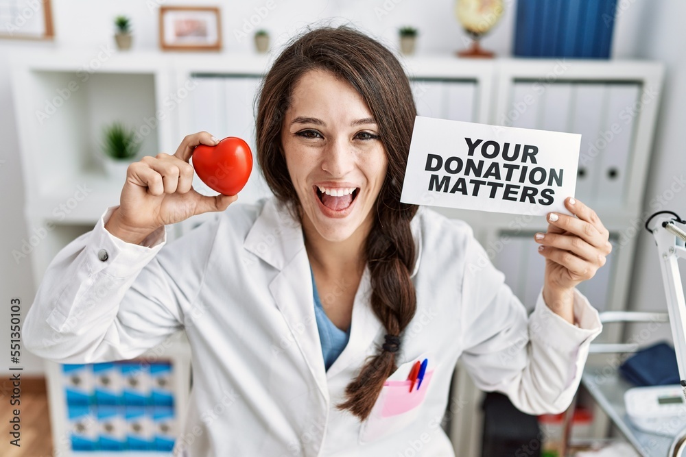 Poster young doctor woman holding your donation matters banner at the clinic smiling and laughing hard out 