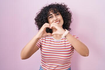 Young middle east woman standing over pink background smiling in love doing heart symbol shape with hands. romantic concept.