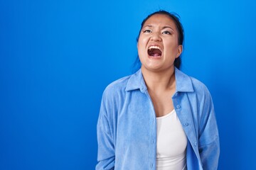 Asian young woman standing over blue background angry and mad screaming frustrated and furious, shouting with anger. rage and aggressive concept.