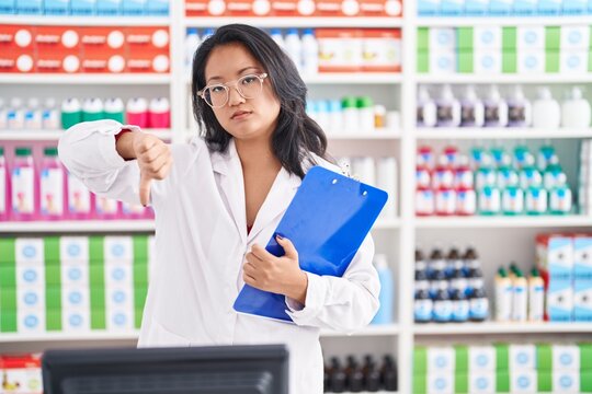 Asian young woman working at pharmacy drugstore holding clipboard with angry face, negative sign showing dislike with thumbs down, rejection concept