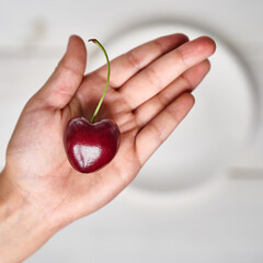 Close-up of a ripe large cherry in the shape of a heart in the hands of a girl, against the background of a white plate. The concept of healthy nutrition and vitamins. High quality photo