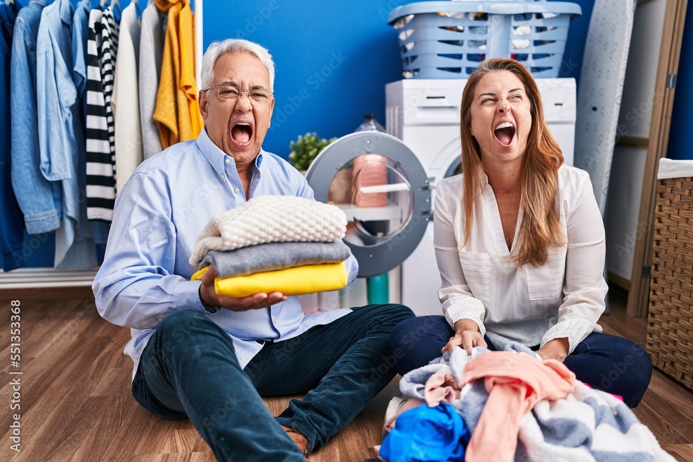 Poster middle age hispanic couple doing laundry sitting on the floor angry and mad screaming frustrated and