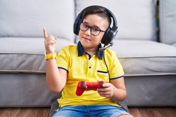 Young hispanic kid playing video game holding controller wearing headphones with a big smile on face, pointing with hand finger to the side looking at the camera. © Krakenimages.com