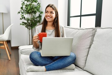 Young latin woman using laptop and drinking coffee sitting on sofa at home