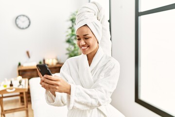 Young latin woman wearing bathrobe using smartphone at beauty center