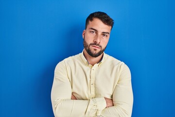 Handsome hispanic man standing over blue background skeptic and nervous, disapproving expression on face with crossed arms. negative person.