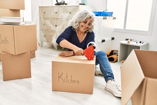 Middle age grey-haired woman smiling happy packing kitchen cardboard box at new home.