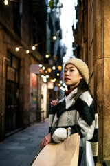 Pensive Chinese female in outerwear and beret with shopping bags in hands standing on street near residential buildings in city