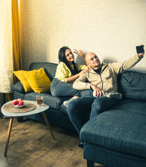 Couple Use Smartphone Device, Sitting on Couch Cozy Apartment. Take selfie together as Boyfriend and Girlfriend. Do video chat on Internet, Use Social Media, Streaming live video