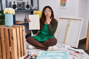 Hispanic young woman holding notebook at art studio covering mouth with hand, shocked and afraid...