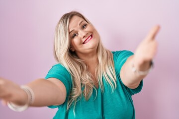 Fototapeta premium Caucasian plus size woman standing over pink background looking at the camera smiling with open arms for hug. cheerful expression embracing happiness.