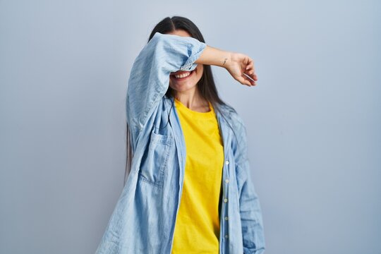 Young hispanic woman standing over blue background covering eyes with arm smiling cheerful and funny. blind concept.