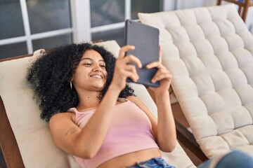 Young hispanic woman using touchpad lying on deck chair at home terrace