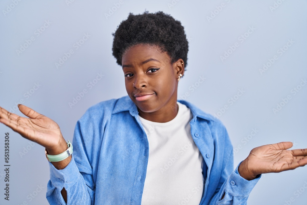 Sticker African american woman standing over blue background clueless and confused expression with arms and hands raised. doubt concept.