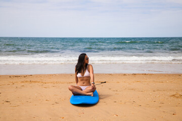 Beautiful young woman sitting on the surfboard on the shore of the beach. The woman is enjoying her trip to a paradise beach. Holiday and travel concept.