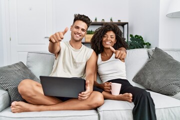 Young interracial couple using laptop at home sitting on the sofa approving doing positive gesture with hand, thumbs up smiling and happy for success. winner gesture.