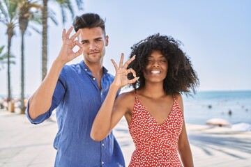 Naklejka premium Young interracial couple outdoors on a sunny day smiling positive doing ok sign with hand and fingers. successful expression.