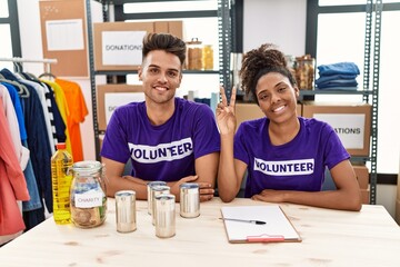 Young interracial people wearing volunteer t shirt at donations stand showing and pointing up with fingers number two while smiling confident and happy.