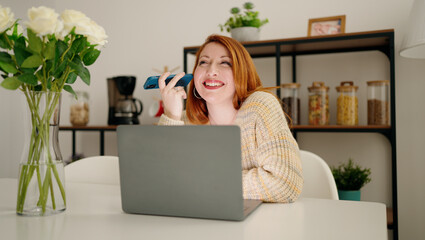 Young redhead woman using laptop listening audio message by smartphone at home