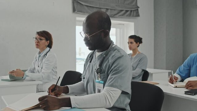 African American general practitioner attending medical training class listening to professor and making notes in notebook