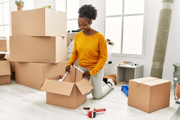 African american woman smiling confident unpacking cardboard box at new home - obrazy, fototapety, plakaty