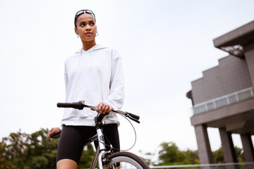 Naklejka premium Low angle view of fit african american woman in sunglasses and hoodie standing near bike outdoors 