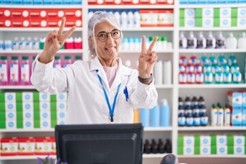 Middle age woman with tattoos working at pharmacy drugstore smiling with tongue out showing fingers of both hands doing victory sign. number two.
