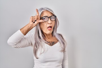 Middle age woman with grey hair standing over white background making fun of people with fingers on forehead doing loser gesture mocking and insulting.