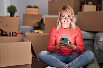 Young blonde woman using smartphone sitting on floor at new home