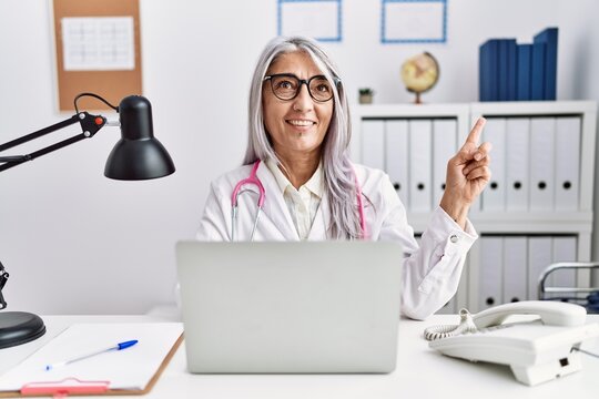 Middle age grey-haired woman wearing doctor uniform working using computer laptop smiling happy pointing with hand and finger to the side