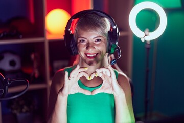 Young blonde woman streamer smiling confident doing heart symbol with hands at gaming room