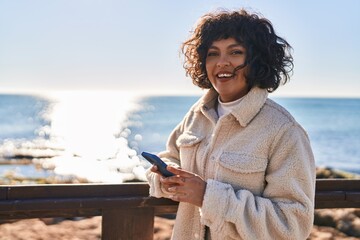 Young beautiful hispanic woman smiling confident using smartphone at seaside