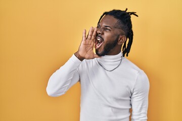 African man with dreadlocks wearing turtleneck sweater over yellow background shouting and screaming loud to side with hand on mouth. communication concept.