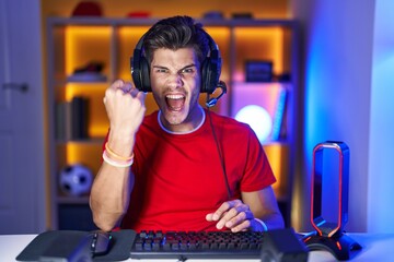 Young hispanic man playing video games angry and mad raising fist frustrated and furious while...