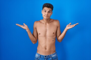 Young hispanic man standing shirtless over blue background clueless and confused expression with...