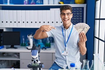 Naklejka na ściany i meble Young hispanic man working at scientist laboratory holding dollars pointing finger to one self smiling happy and proud
