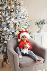 Vertical photo of funny little boy in santa clause costume opening his christmas present sitting in a chair near the christmas tree at home